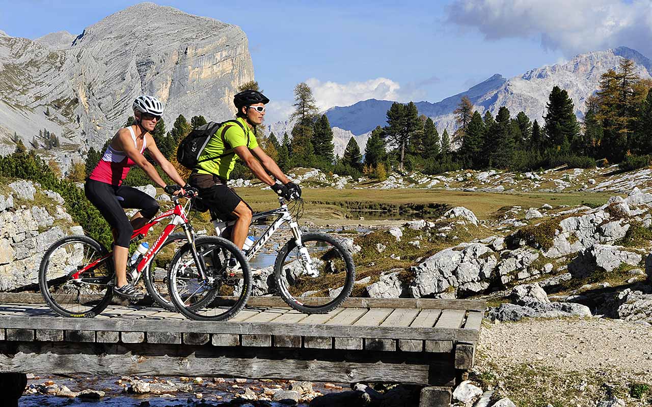 Two boys on a bike cross a wooden bridge and in the background the Dolomites